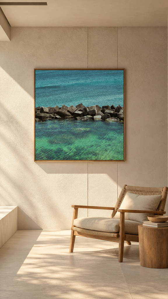 Square fine art photograph of a stone breakwater in turquoise water, in a light wood float frame, hung at eye level on a beige plaster wall beside a rattan armchair